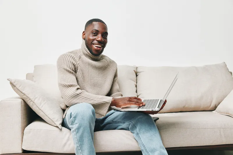 Happy Black man working on a laptop, wearing a cozy sweater, sitting on a sofa in a bright, minimalist living room with a neutral color palette