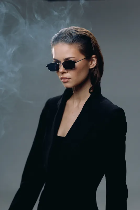 Stylish woman wearing sunglasses and a black outfit with smoky background and serious expression.