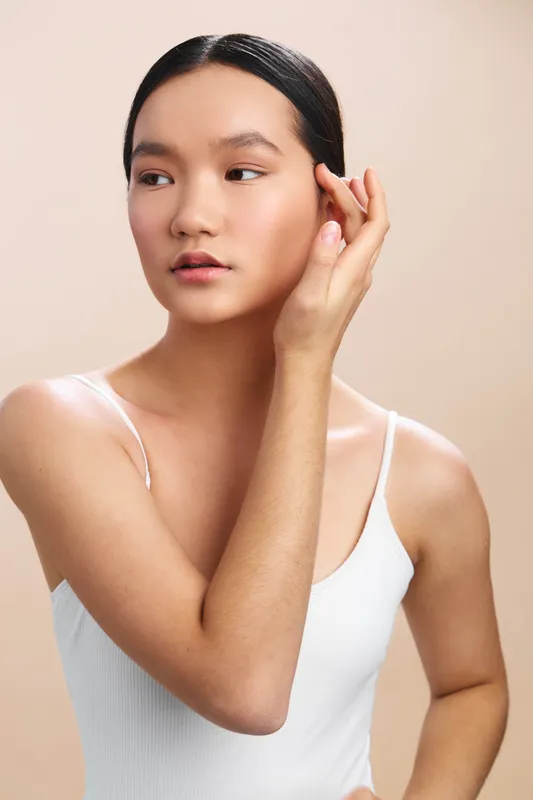 asian woman in a white tank top adjusts her hair in a soft studio setting, creating a calm, elegant portrait with gentle lighting and neutral background.