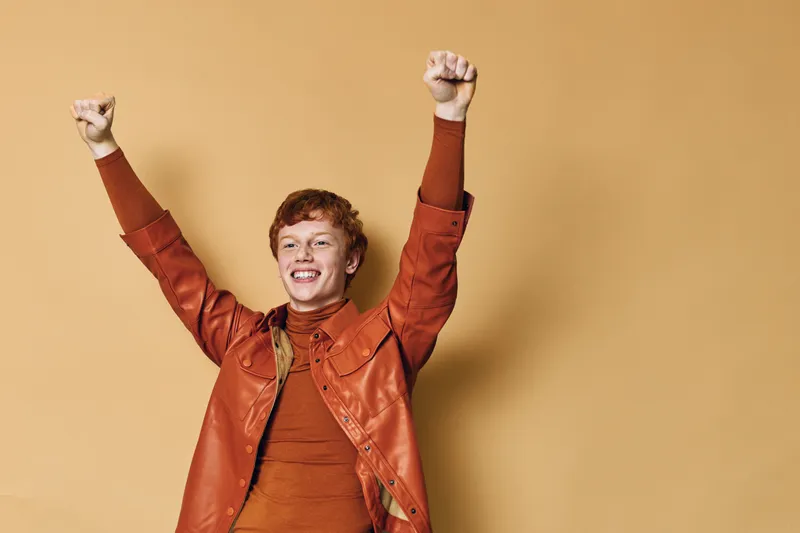 Happy young man celebrating with raised hands wearing brown leather jacket and turtleneck sweater on beige background. Excited male with red hair expressing joy and success.