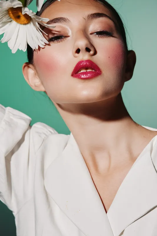 Fashion model with glossy red lips and rosy blush poses against teal background wearing white and a daisy flower near face, spotlighting bold beauty and creative styling.