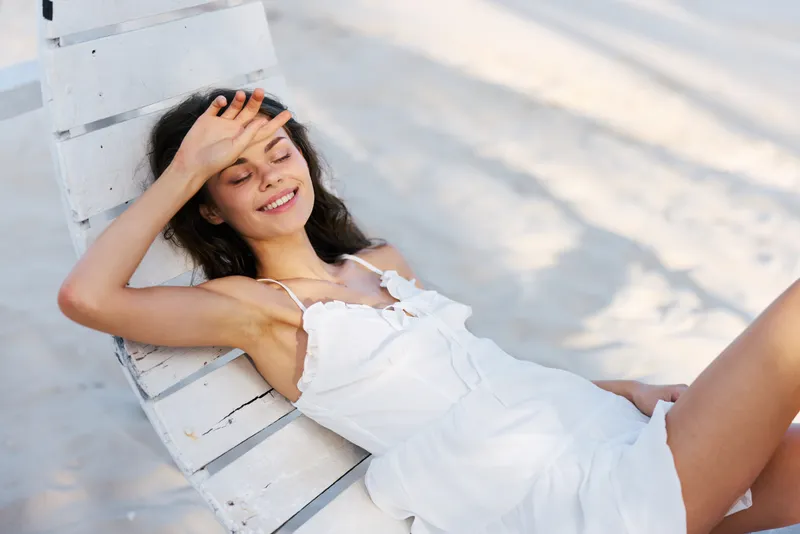 Young woman relaxing on a sunlit beach chair, smiling with eyes closed, enjoying warm breeze and seaside calm, wearing a light white dress, carefree vacation mood and sunlit serenity.