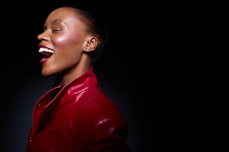 Happy black African woman with dark skin laughing in red leather jacket on black background. Studio portrait showing joy, fashion, and confident positive mood of young female model.