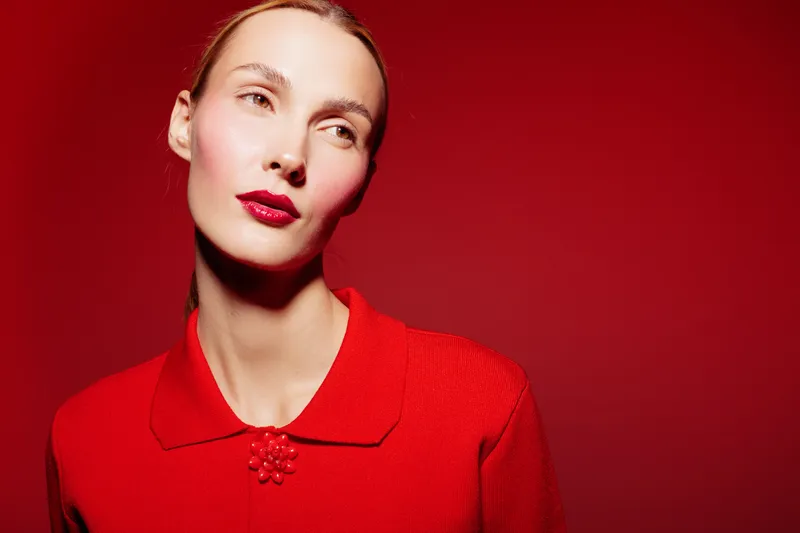 Portrait of a woman in red sweater with glossy red lips on red background. Studio lighting highlights her smooth skin and elegant expression. Concept of fashion and beauty.