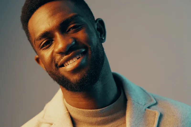 Smiling African man wearing a light jacket with a modern hairstyle, expressing confidence and joy against a gradient background, conveying a sense of positivity and style