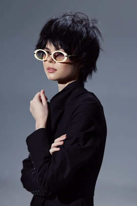 Stylish woman wearing white sunglasses and black outfit posing indoors with confident expression and modern haircut