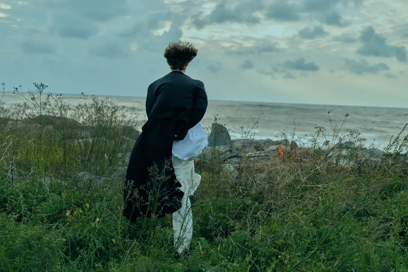 Back view of a figure standing on a windy coastal hillside, gazing toward the sea, wearing a dark coat and white garment, creating a moody, lone silhouette overlooking waves.