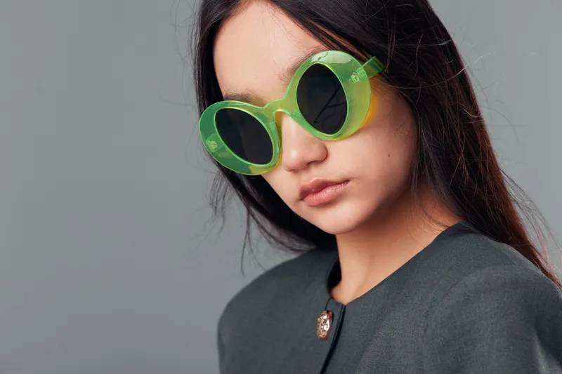 Woman with long dark hair wears oversized round green sunglasses and gray coat posing against plain background for fashion style portrait and eyewear trend in studio.