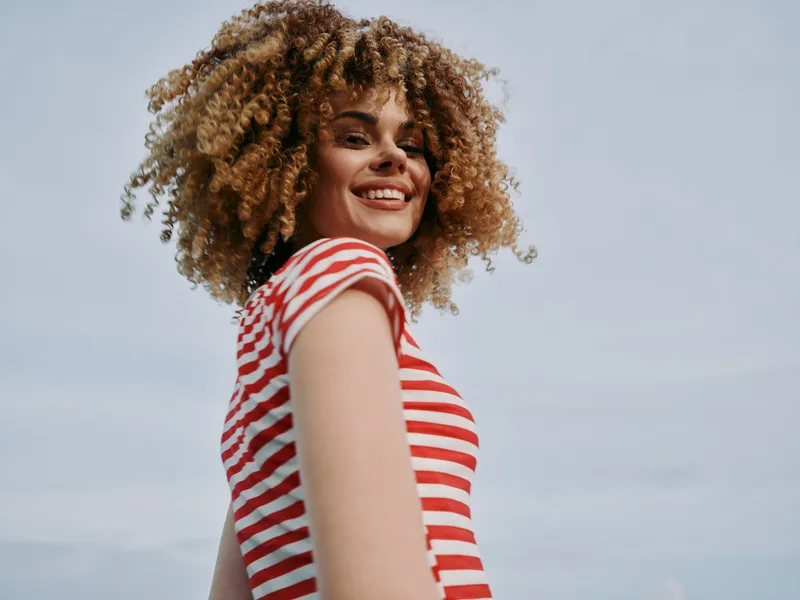 Portrait of a smiling woman with curly hair in a red and white striped tee, standing outdoors under a clear blue sky, radiating carefree confidence and summer warmth.