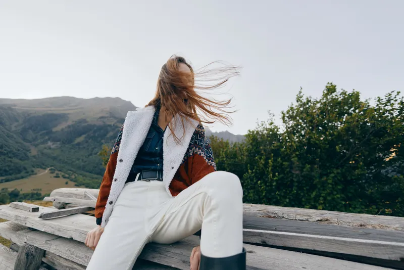 Woman sitting on wooden bench in mountains, hair blown by wind across her face, wearing cozy jacket, white pants and boots, relaxed outdoors travel portrait in natural landscape.