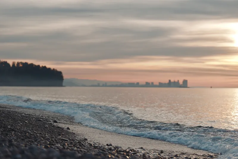 Beach at sunset with calm sea, pebbled shoreline and distant city skyline on the horizon, gentle waves and warm light creating a tranquil coastal scene.