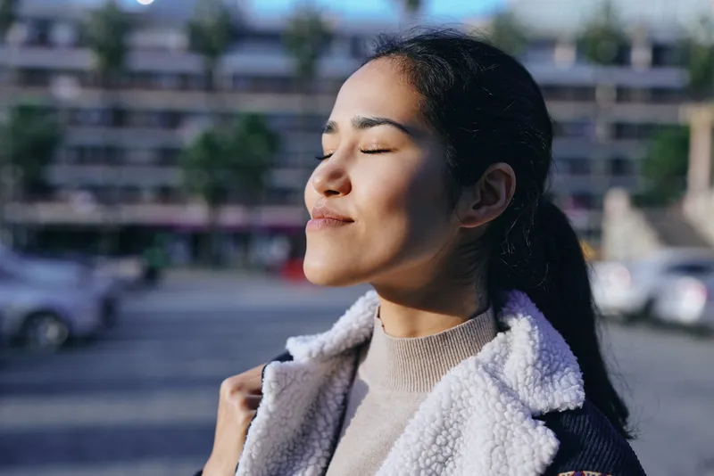 Sunlit outdoor scene with a calm woman closing her eyes, wearing a cozy jacket and sweater, warm daylight on her face, relaxed mood, tranquil moment, casual style, serene atmosphere