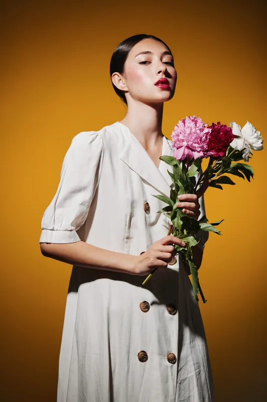 Young woman with glossy black hair wears a white dress while holding colorful flowers. Shot on orange background. Concept for creative beauty and fashion editorial.