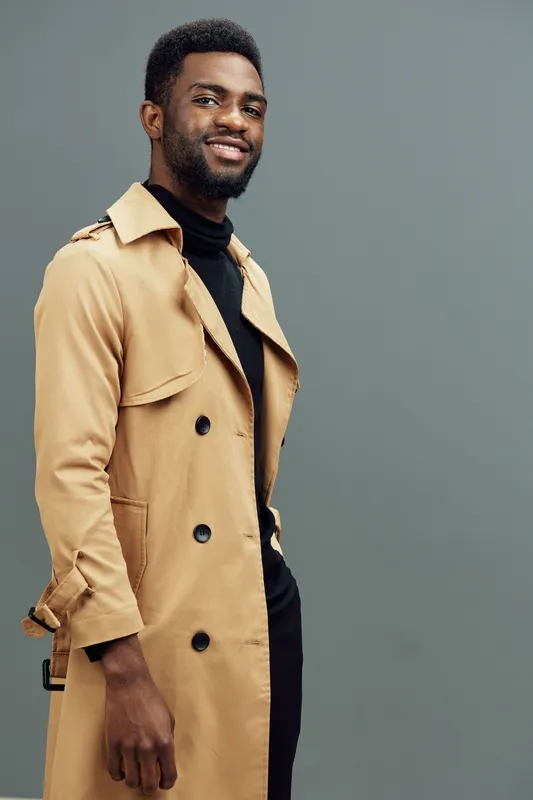 Stylish African American man in a beige trench coat, smiling confidently against a soft gray background, exuding modern fashion and charisma