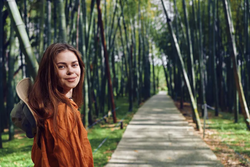 Woman with backpack on path through bamboo forest, travel portrait of a smiling young woman looking over her shoulder on a paved walkway, nature trip and outdoor exploration.