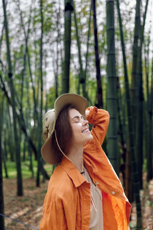 Woman in a bamboo forest wearing a straw hat and orange jacket, portrait shot with a bright smile, enjoying nature outdoors with green trees, sunlight and peaceful travel vibes.