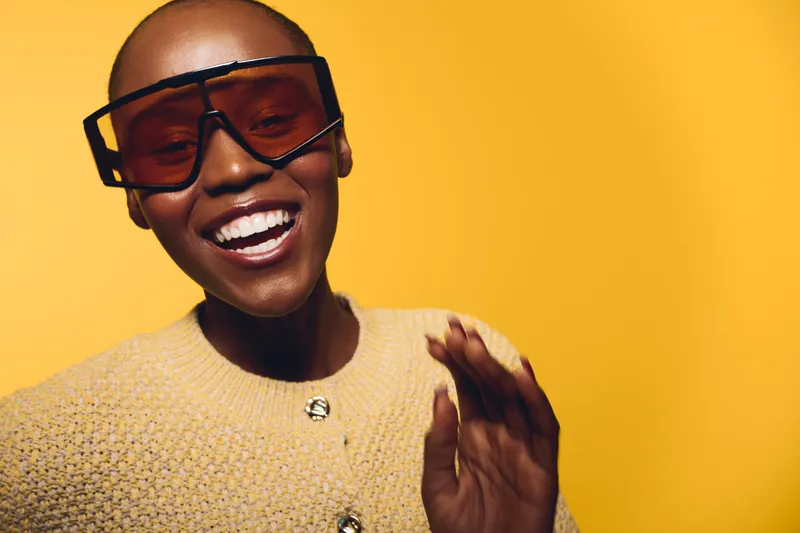 Happy Afro American woman with dark skin wearing large sunglasses and beige sweater laughs joyfully on yellow background. Concept of fashion, confidence, and positive mood in studio portrait.
