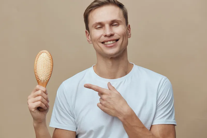 Smiling man holding hairbrush pointing to his head on beige background, casual clothing, cheerful expression, studio portrait, people lifestyle concept