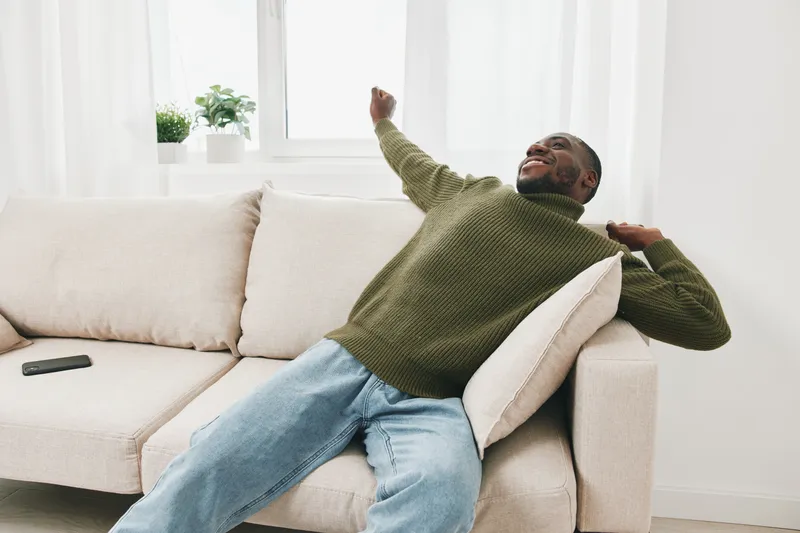 Young Black man enjoying a moment of relaxation on a light colored couch in a cozy, modern living space with a soft green sweater and denim jeans