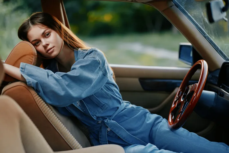 Young woman in denim outfit relaxed in vintage car, capturing a serene mood in nature with soft colors. Emphasizes tranquility, youth, and adventure.