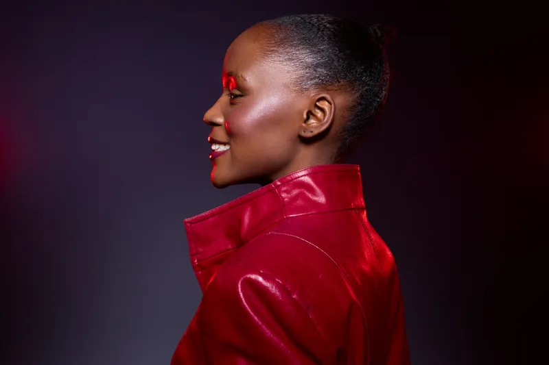 Profile of a smiling African woman with dark skin wearing a shiny red leather jacket. Studio portrait with dramatic lighting on black background. Concept of fashion, confidence, and style.