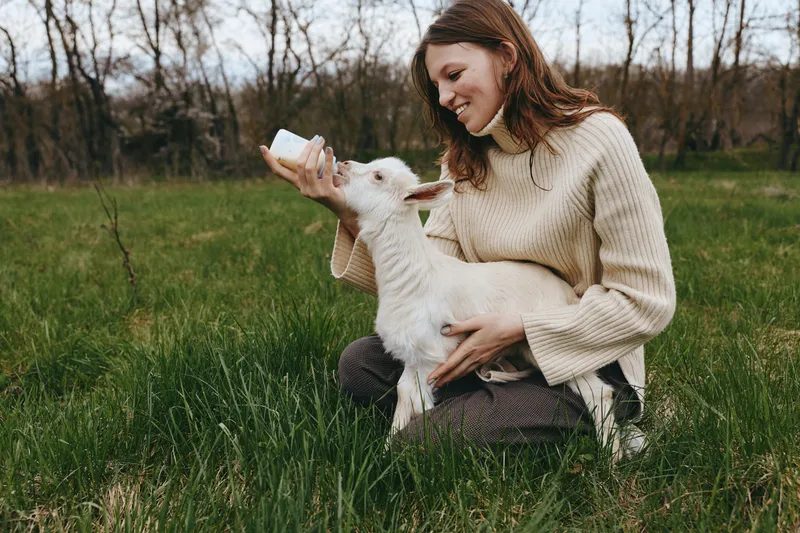 Cheerful woman outdoors, feeding a kid goat with a bottle, peaceful countryside setting, soft sweater, natural colors, warm light, joyful connection, relaxed posture, candid lifestyle moment
