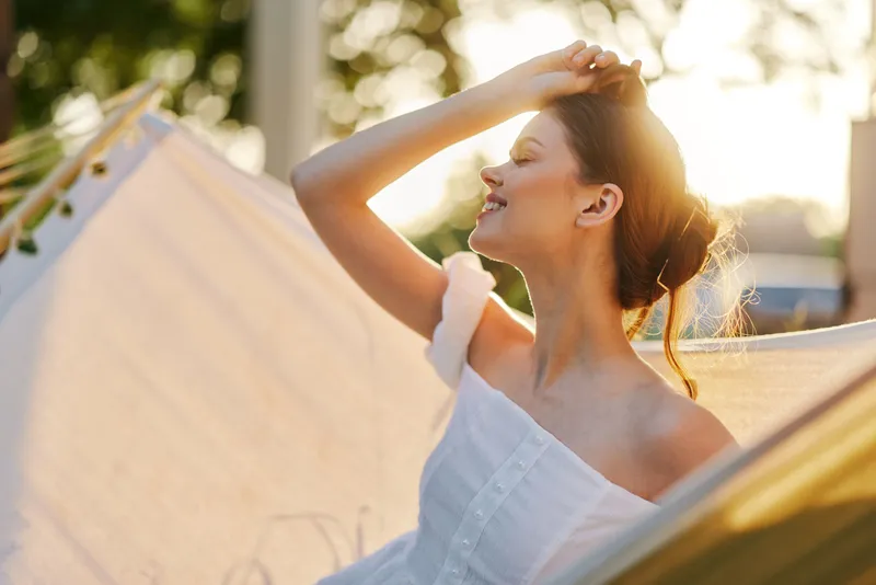 Joyful young woman enjoying a sunny day outdoors, wearing a white dress, with a soft smile, surrounded by a bright, natural background