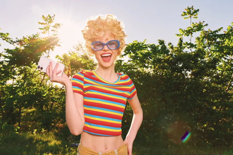 A woman in a colorful striped crop top stands outdoors under sunny light, laughing with sunglasses. A retro film color palette enhances a carefree lifestyle and outdoor happiness.