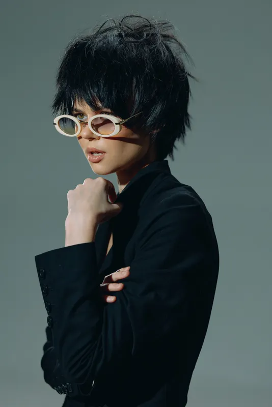 Stylish woman with short black hair and white sunglasses wearing a black jacket with a moody gray background