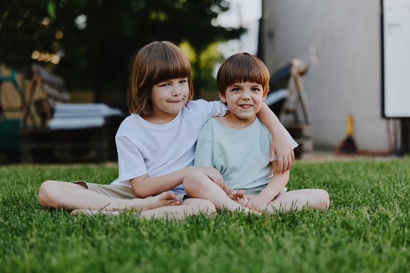 Two cheerful children sitting on green grass, enjoying a sunny day, displaying sibling affection and happiness, with a soft natural background.