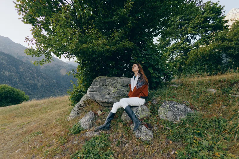 Woman sitting on rocks under a large tree in mountains, countryside nature scene with boots and red jacket, relaxed portrait of outdoor travel, serene meadow and greenery.