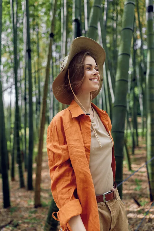 Woman smiling in hat among bamboo forest, portrait of traveler in nature wearing orange shirt and casual outfit, peaceful outdoor scene for eco travel and adventure lifestyle.