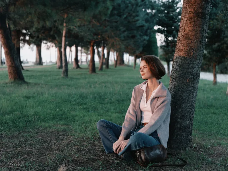 A woman sits beside a tree in a peaceful park, gazing into the distance with a calm expression, enjoying a quiet moment amid green grass and gentle light.
