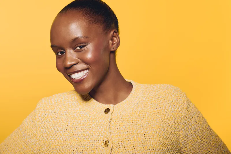Smiling African woman with dark skin wearing a yellow knit sweater poses against yellow background. Concept of happiness, confidence, and natural beauty in fashion portrait.