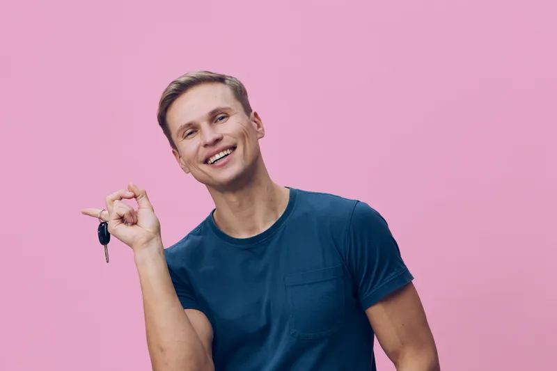 Smiling young caucasian man hold keys in hand wear blue t-shirt look camera isolated pink background concept digital nomad lifestyle and remote work with freedom and joy