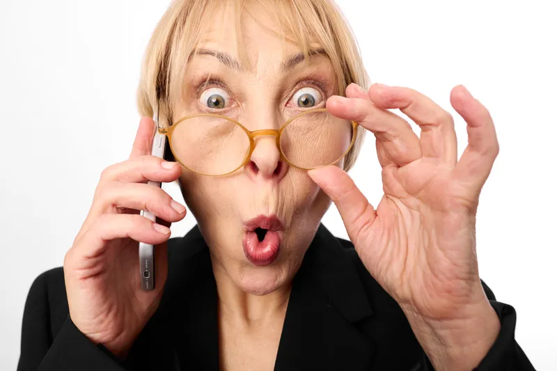 Surprised business woman with glasses holds phone, hands raised near face, shocked open mouth, isolated on white background portrait, modern communication moment.