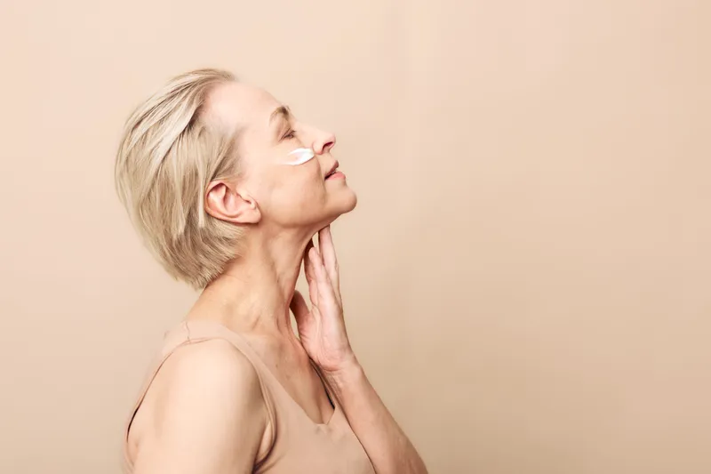 Mature woman with short blonde hair applies moisturizing cream on her cheek, eyes closed, and gently touches neck. Skincare concept for aging skin, beauty routine, and self-care in studio.