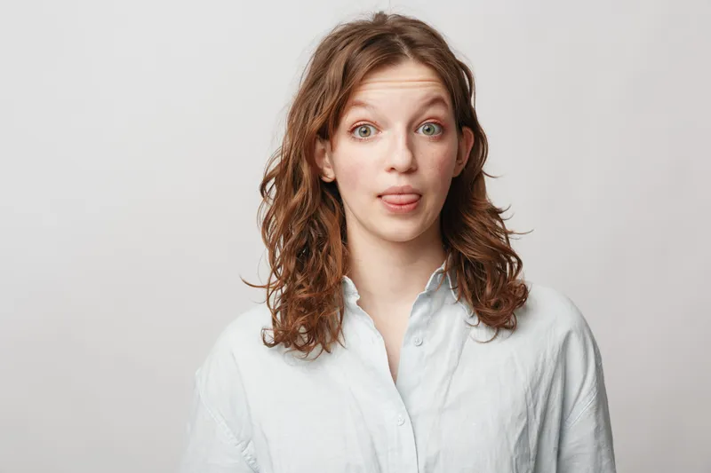 Young woman with curly hair wearing white shirt makes funny face sticking out tongue. Studio portrait on light background. Concept of humor, playfulness, and casual lifestyle.