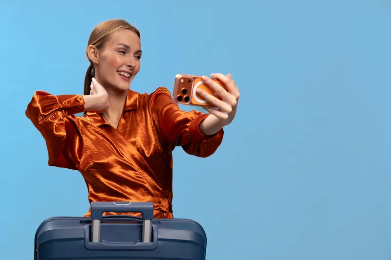 Happy woman in shiny orange blouse takes selfie with smartphone while holding handle of blue suitcase against blue background. Travel concept for vacation, journey, and lifestyle.