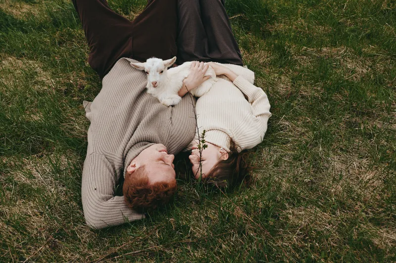 Couple enjoying a relaxed lifestyle outdoors, cuddling on the grass with a tiny goat kid, capturing tender romance and affection in a natural pastoral setting, warm sunlit moment for couples and