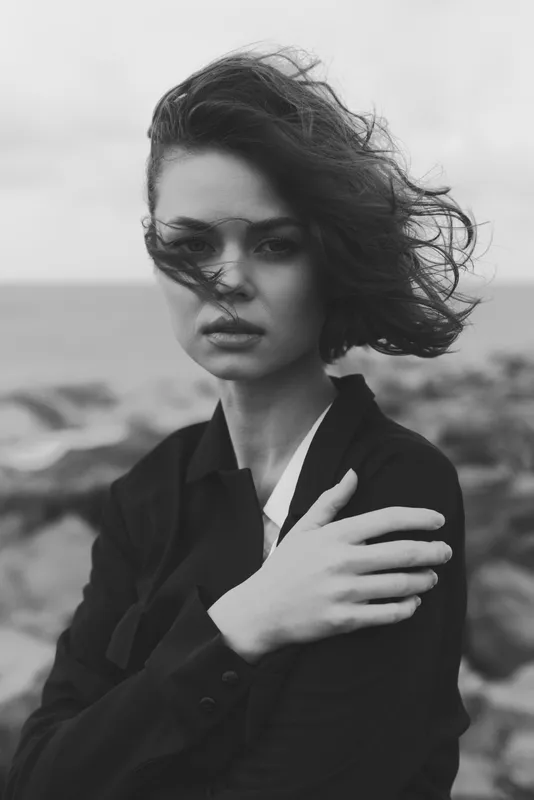 Moody wind swept black and white image of a woman on a windy seaside, fashion inspired pose, thoughtful expression, outdoors by the coast and rocks