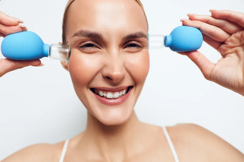 Smiling woman using blue facial suction cups on cheeks. Close-up with white background. Concept of skincare, facial massage, anti-aging, and beauty routine for healthy skin.