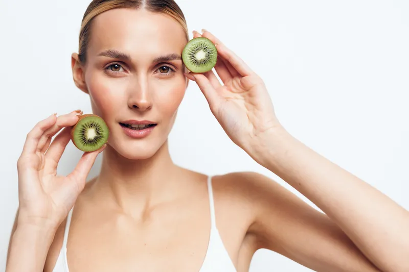 Young woman holds two halves of fresh kiwi fruit near face with natural skin expression isolated on white background with healthy lifestyle concept and beauty care.