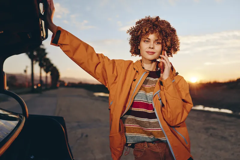 Woman lifestyle outdoors at sunset, curly hair smiling near car, orange jacket casual fashion, relaxed pose with arm raised by parked vehicle, golden sky light