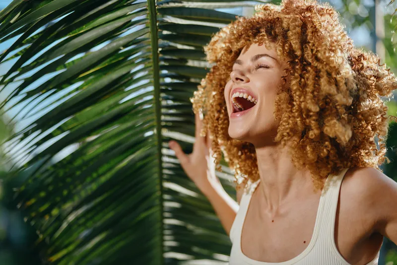 joyful woman with curly hair laughing outdoors near tropical palm leaves in bright sunlight, expressing happiness and natural beauty in summer atmosphere.