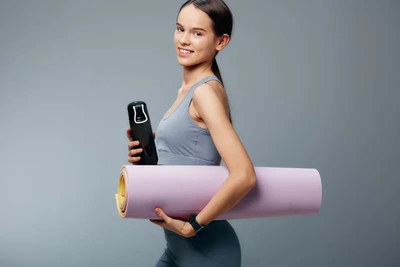 Fitness woman in athletic wear carries a yoga mat and water bottle, ready for gym routine and training, showcasing a focused sport vibe in a clean studio.