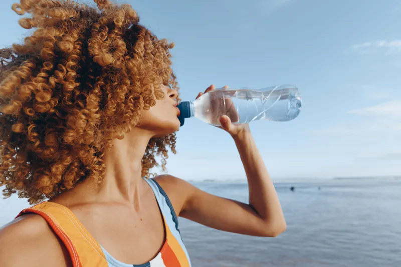 Woman with curly hair drinking water from clear plastic bottle beside the ocean, sunny summer outdoors, hydration and refreshment concept, health beach lifestyle, rehydration
