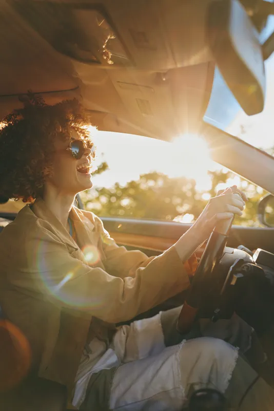 Woman driving a car, sunlight glare, retro film color, lifestyle scene, candid smile, joyful moment, portrait of a carefree traveler, warm tones.
