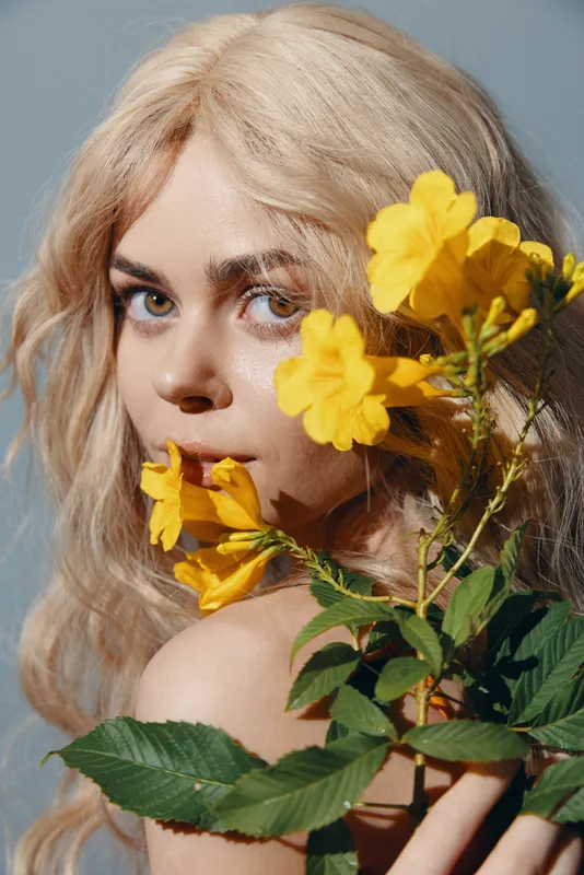 Безмятежная молодая женщина с blond-волосами держит яркие желтые цветы, создавая потрясающий контраст на мягком голубом фоне, отражая красоту и спокойствие