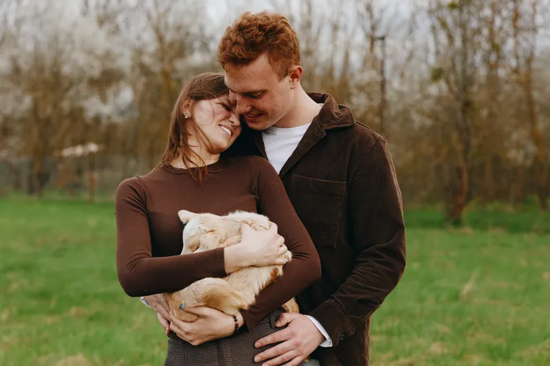 A loving couple shares a tender moment outdoors, holding a goat kid in a grassy park. Warm light and gentle smiles convey connection, care, and simple happiness in a countryside lifestyle.